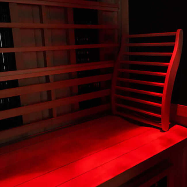 Inside Finnmark Full Spectrum Infrared wooden sauna, a bench, a slatted backrest, illuminated by strong red ambient light inside a dark room and visible Spectrum Plus and Carbon 360 infrared heater panels.