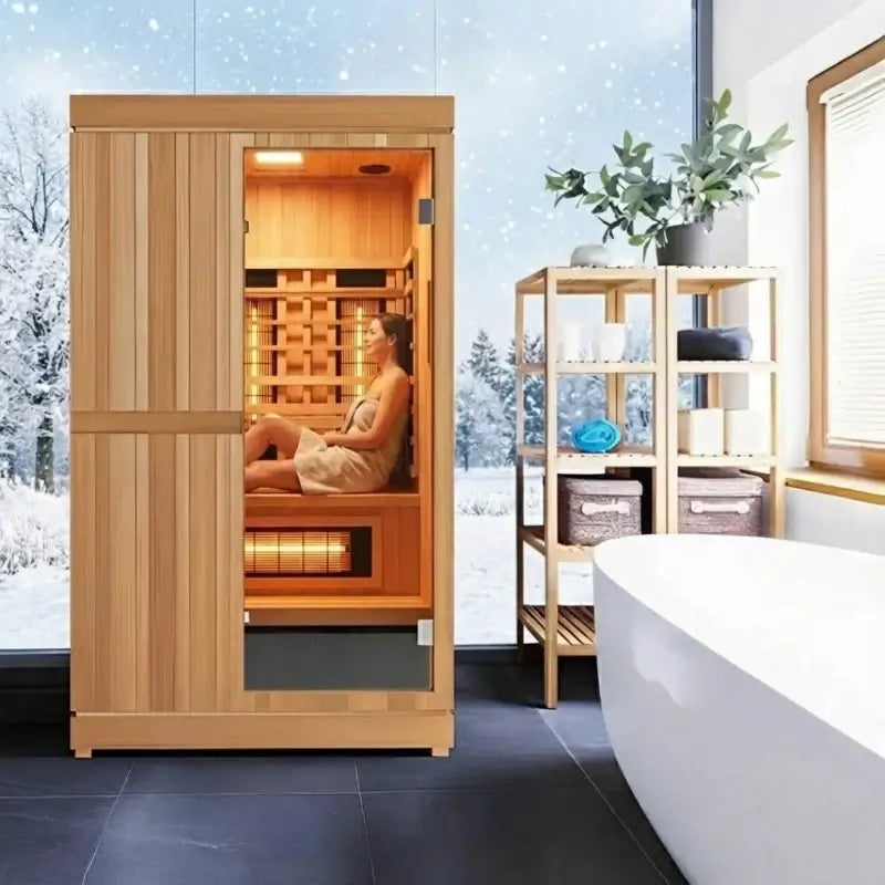 Finnmark FD-4 wooden hybrid sauna room with a glass door, shown with a woman sitting inside. The room is styled in a modern bathroom setting with a large window showing a snowy outdoor view, a white bathtub, and a wooden shelf with spa accessories.