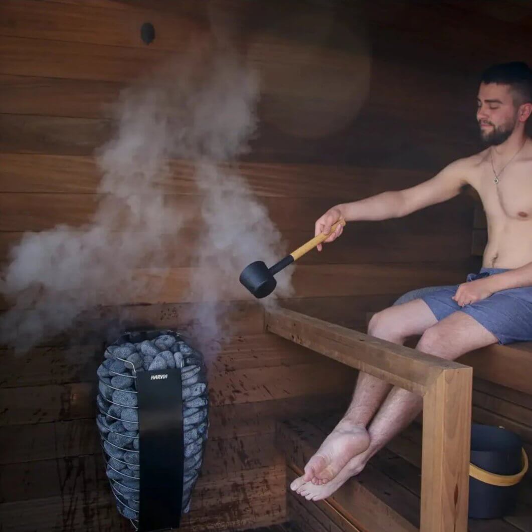 Person sitting in a sauna using a ladle to pour water on Harvia Spirit electric sauna heater that is emitting steam.