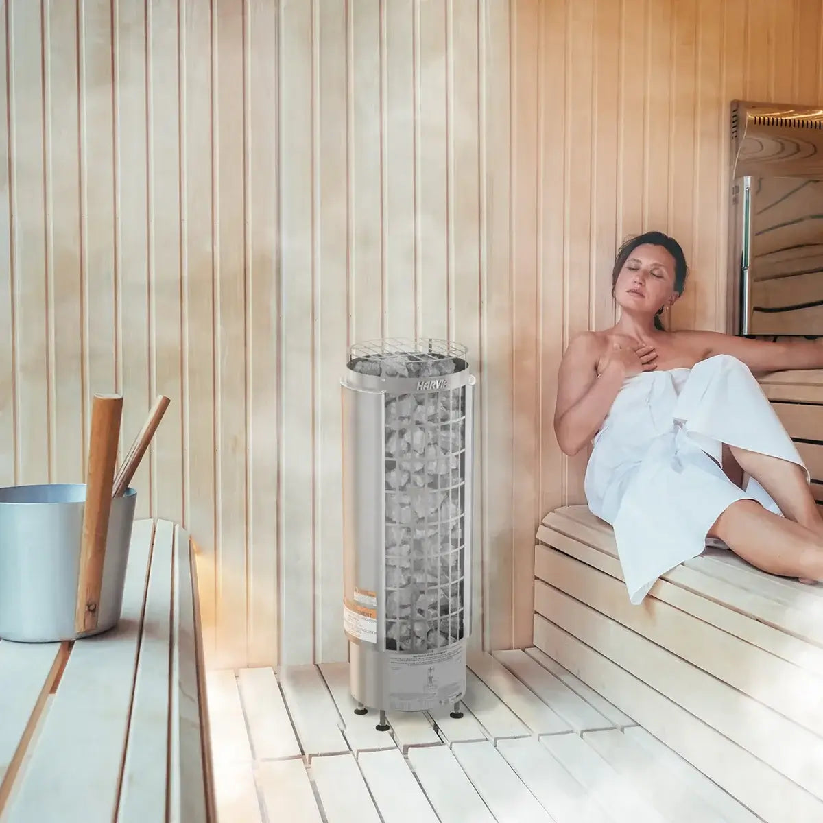 Woman in a sauna with a Harvia Cilindro Half Series heater surrounded by wooden walls and floor.
