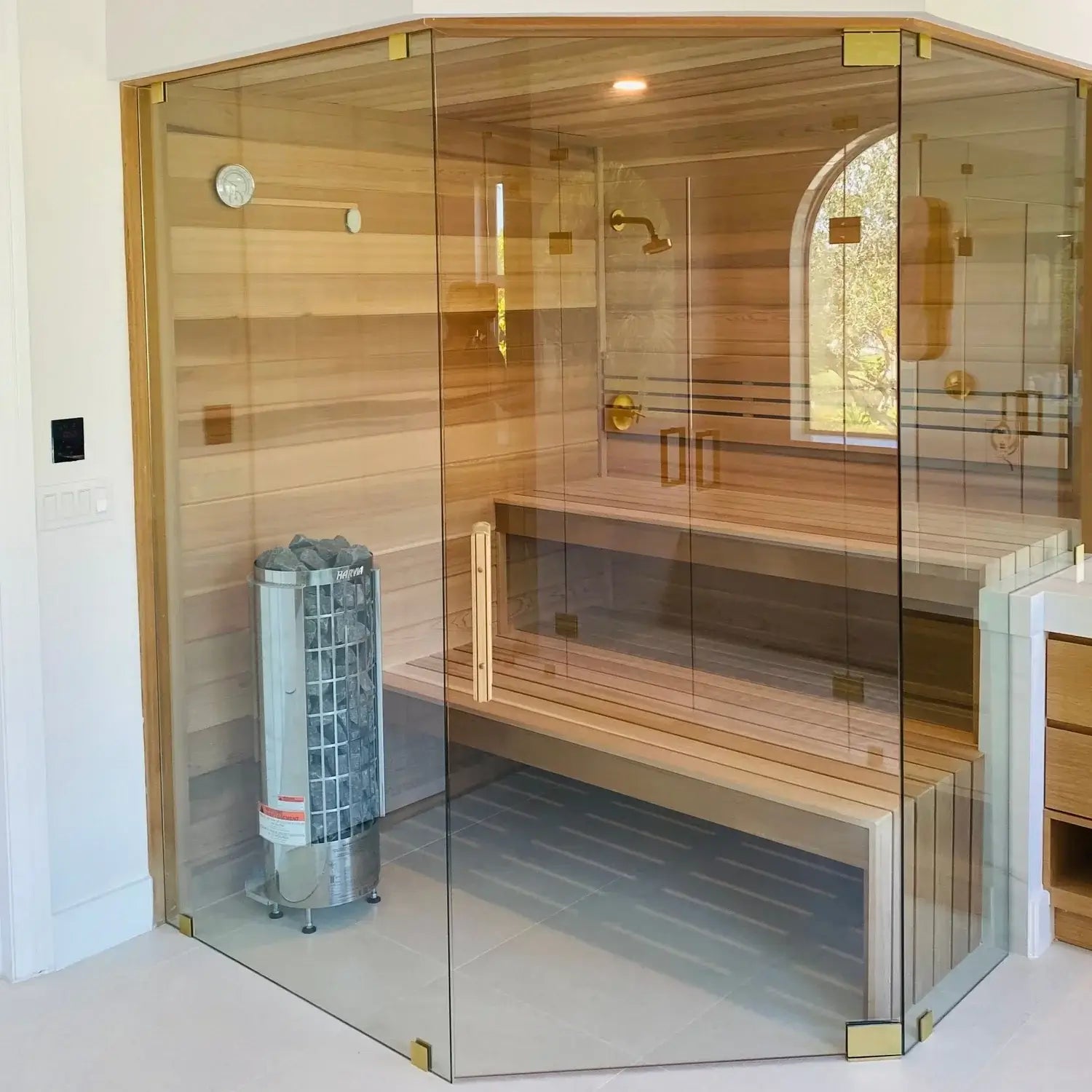 A corner glass sauna room enclosure with a light wood interior, double wooden benches and a gray Harvia Cilindro electric sauna heater placed in one of it's corners