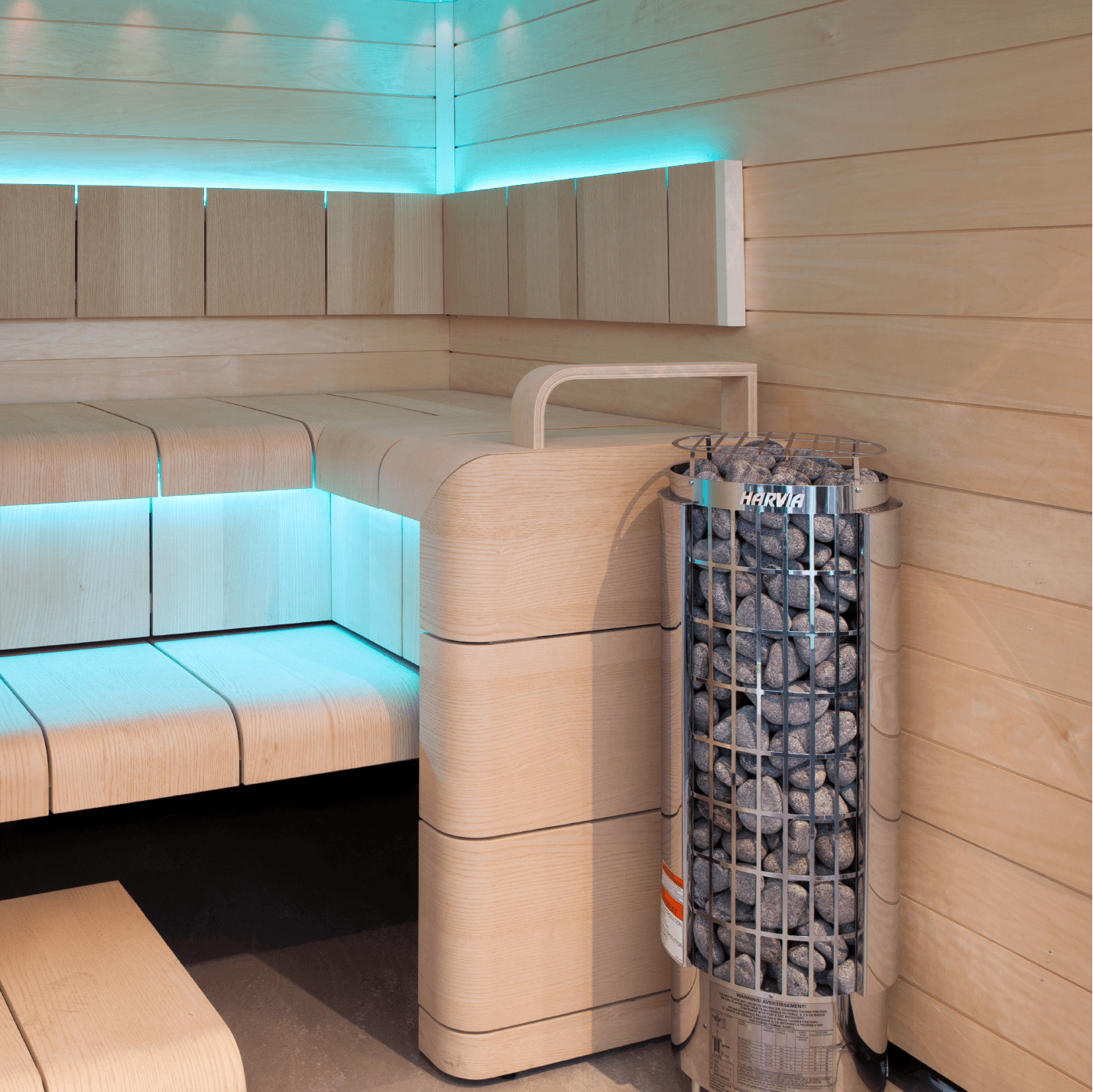 Modern wooden sauna interior with tiered seating, glowing blue LED lighting, and a tall stone-filled cylinder-shaped Harvia Cilindro Half Series Electric Sauna Heater, beside the bench area.