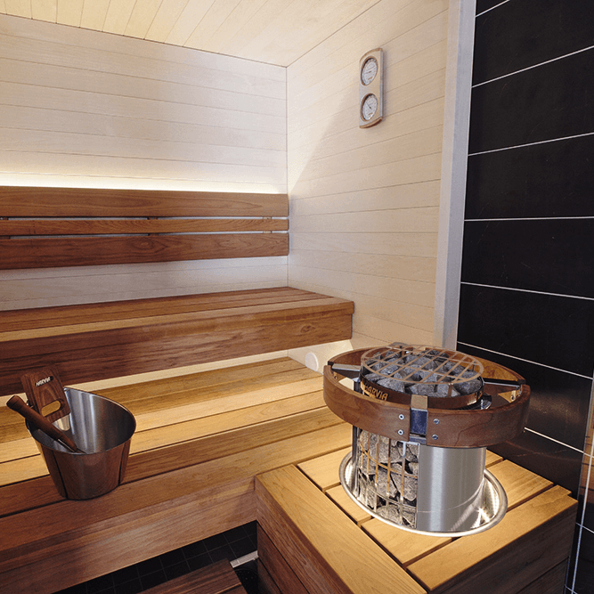 A wooden sauna interior with tiered wooden benches, a metal ice bucket, and a Harvia Cilindro stainless steel heater integrated through a sauna bench with an embedding flange, containing dark heated stones. The walls are light wood paneling with a dark tiled section on the right.