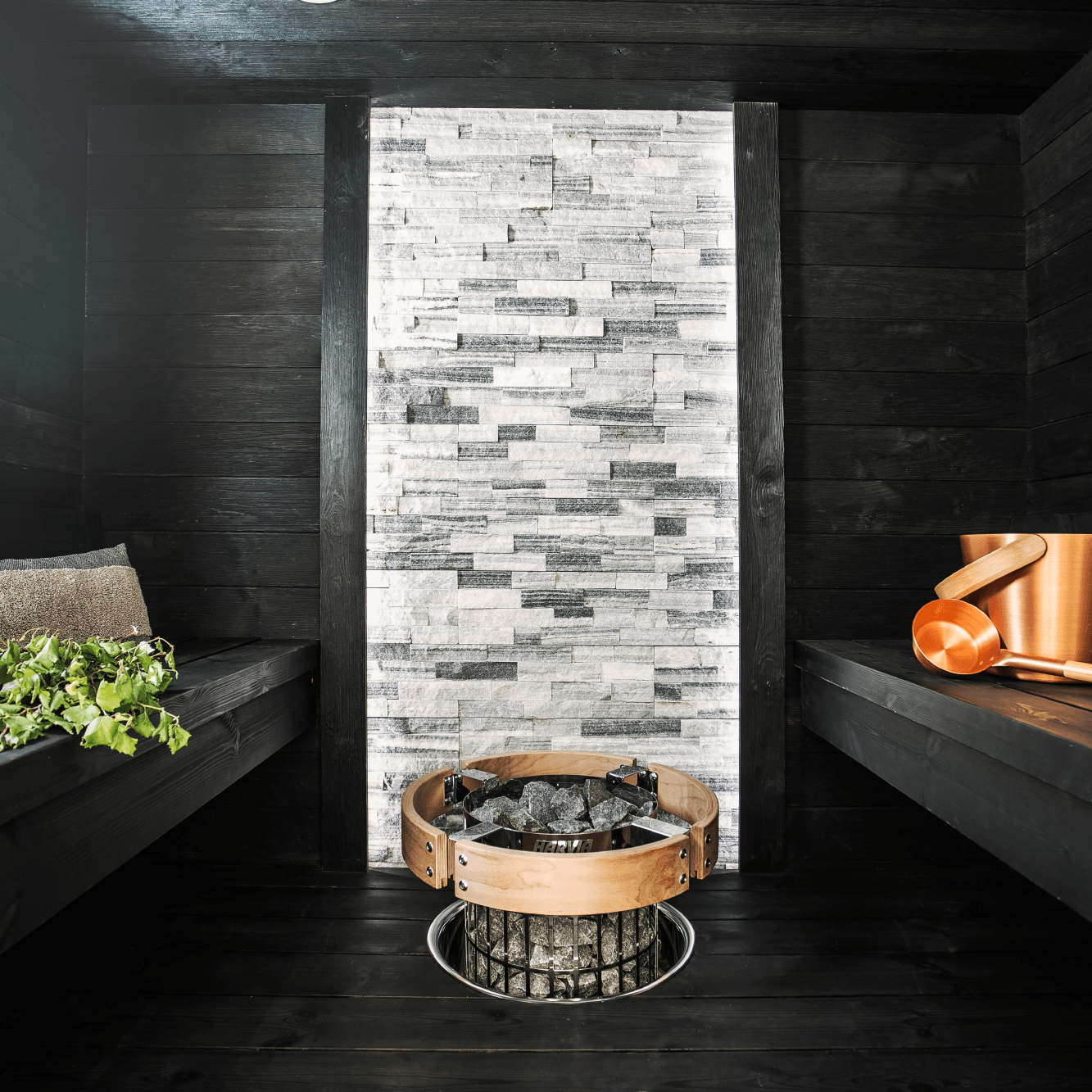 A modern sauna interior with dark wood walls and benches, featuring a central stone-tile accent wall and a round Harvia Cilindro Half Series Electric Sauna Heater with black rocks in the foreground.
