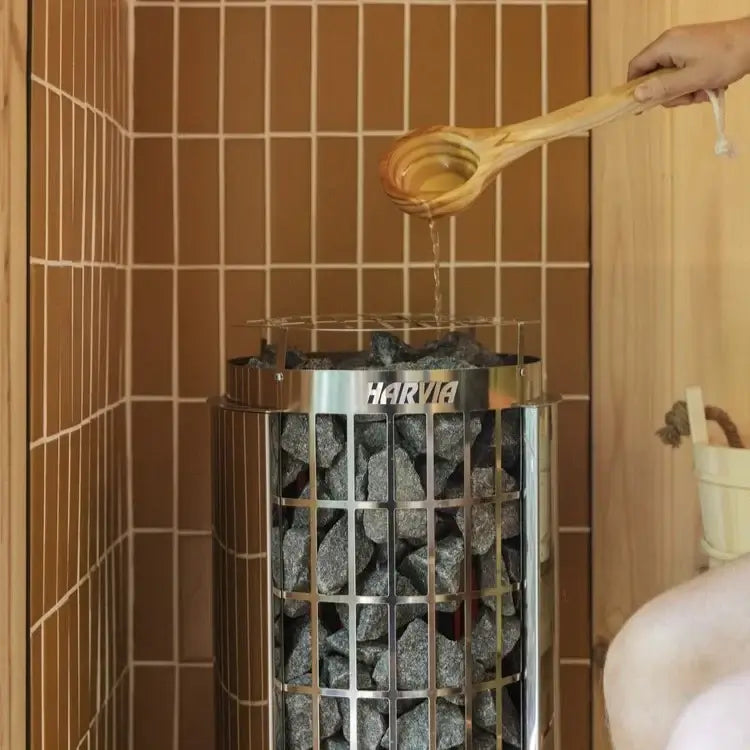 A Harvia Cilindro sauna stone heater filled with dark heating stones, with a person pouring hot water from a wooden ladle. The scene is set inside a wooden sauna with brown tiled walls.