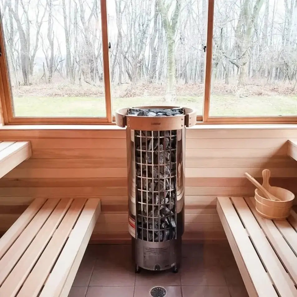 A freestanding cylindrical Harvia Cilindro half series electric sauna heater with exposed stone pillar, placed in the center of a wooden sauna interior with benches and a window showing a wooded outdoor scene.