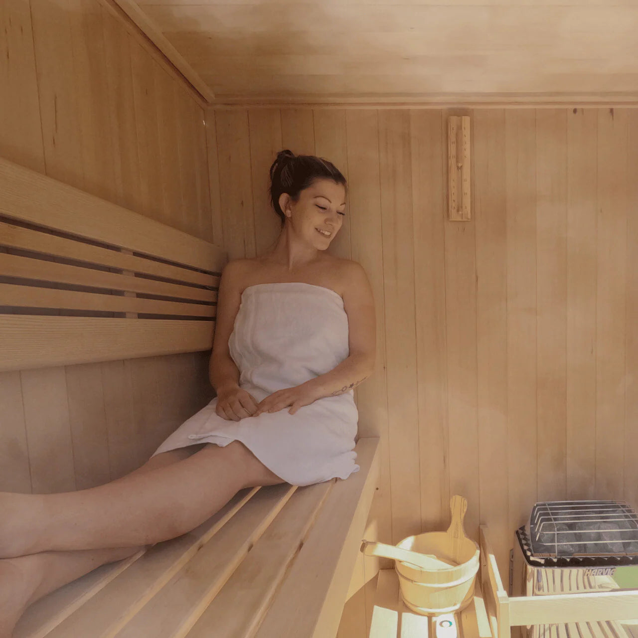 Woman sitting in a wooden sauna with a towel wrapped around her looking at her Harvia KIP sauna heater.