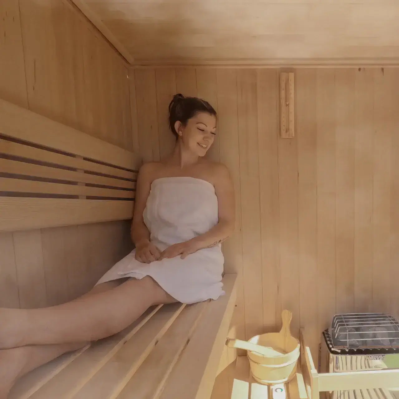 Woman sitting in a wooden sauna with a towel wrapped around her looking at her Harvia KIP sauna heater.