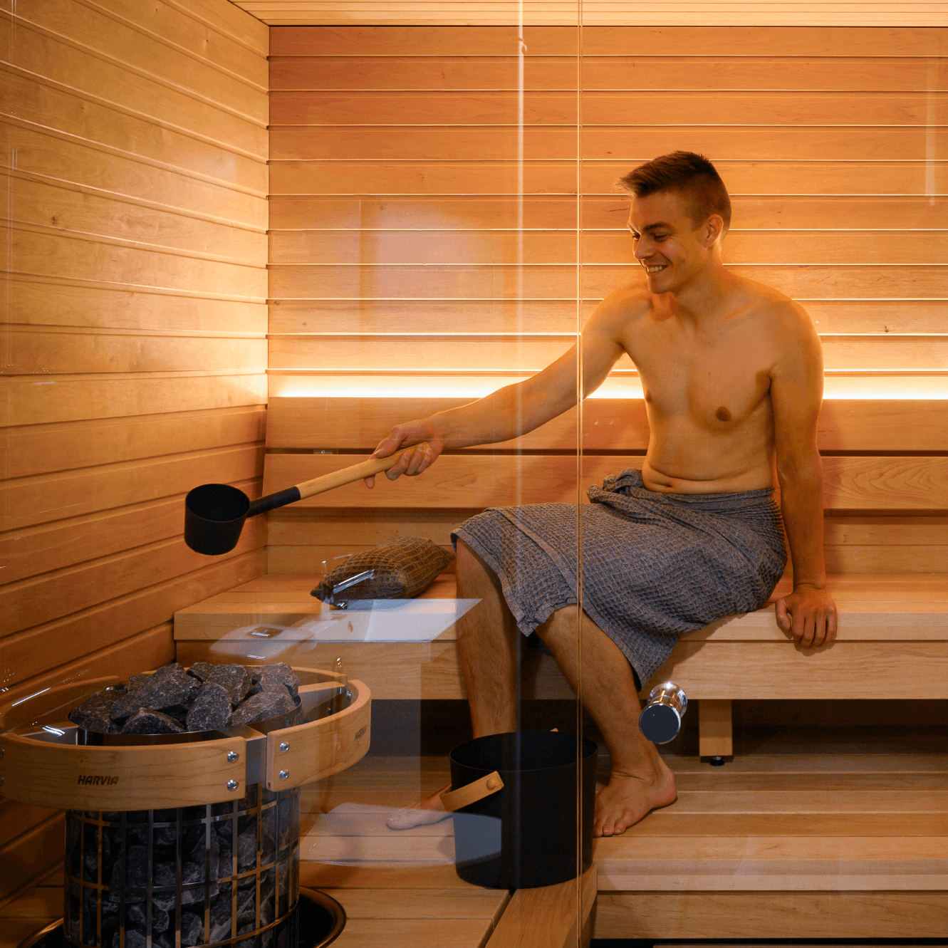 A shirtless man wrapped in a gray patterned towel, sitting on wooden sauna benches inside a wooden sauna, holding a ladle over a Harvia Cilindro half series electric sauna heater with exposed stone pillar