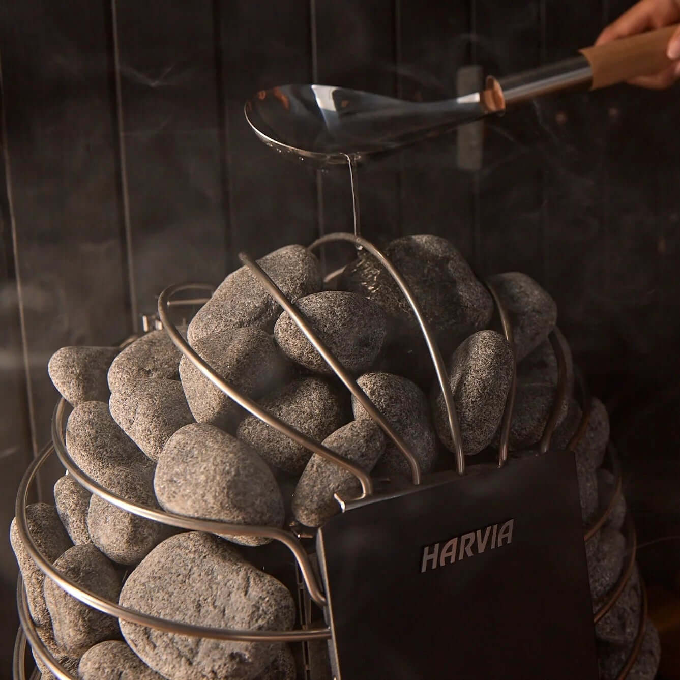 Stainless steel Harvia Spirit electric sauna heater with stones, and a hand holding a ladle and pouring water on the heater, on a dark background