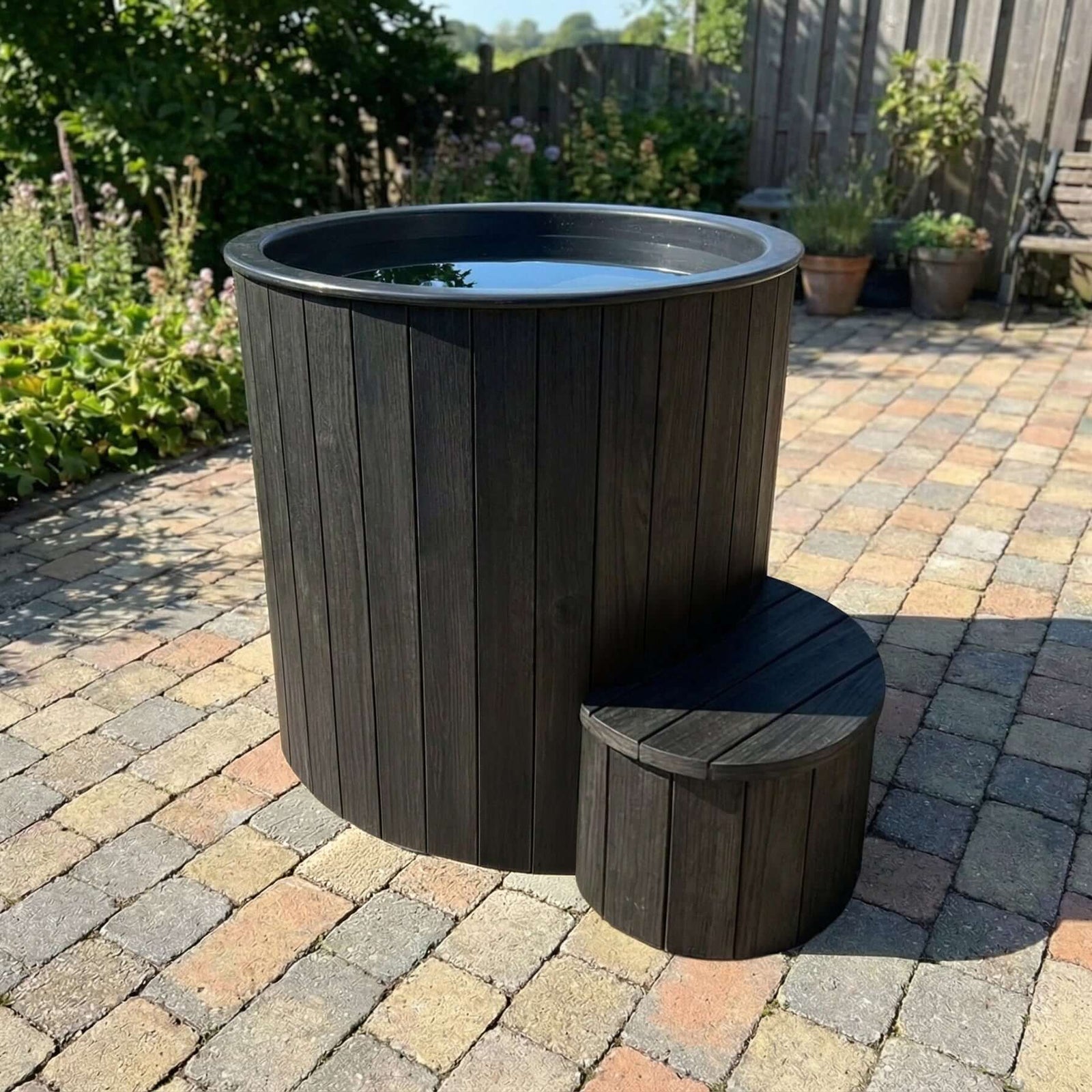 SaunaLife's dark charcoal gray outdoor S1B hot and cold tub with a matching curved step stool, set on a paved backyard patio with garden plants and a wooden fence in the background.