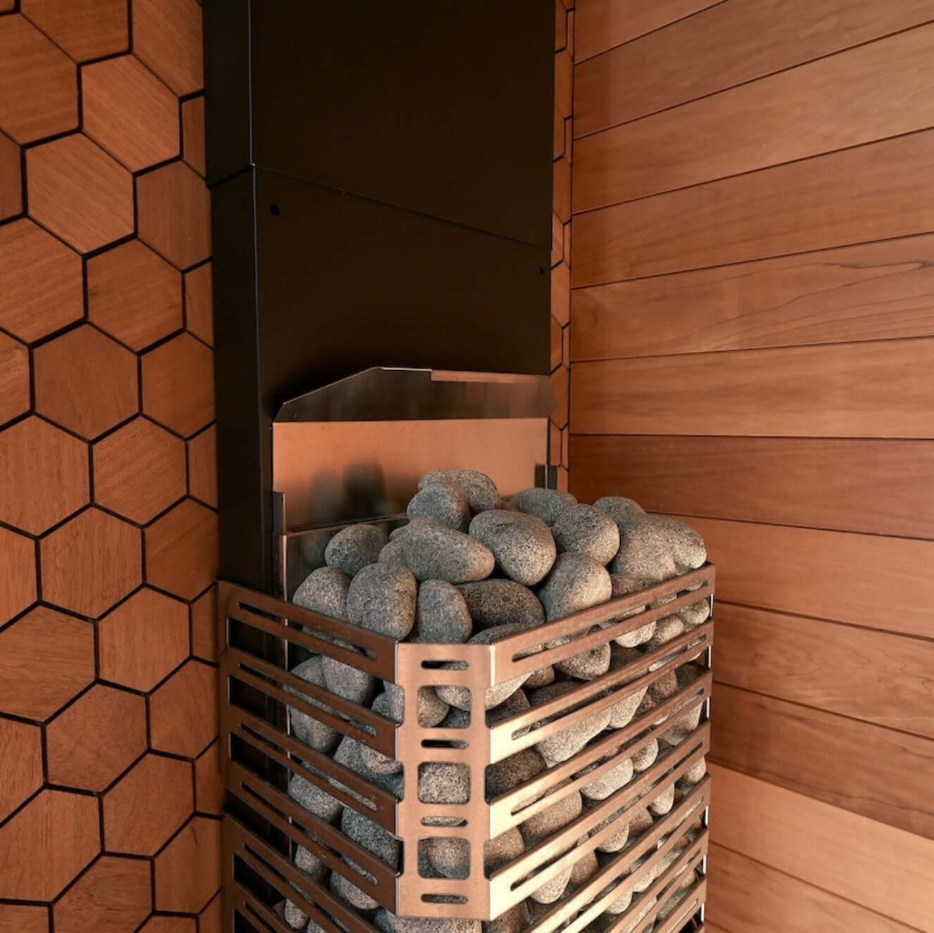 A modern Saunum AIR 5 stainless steel sauna heater with open stone basket filled with gray stones, set against a background of warm wood paneling and hexagonal tiles.