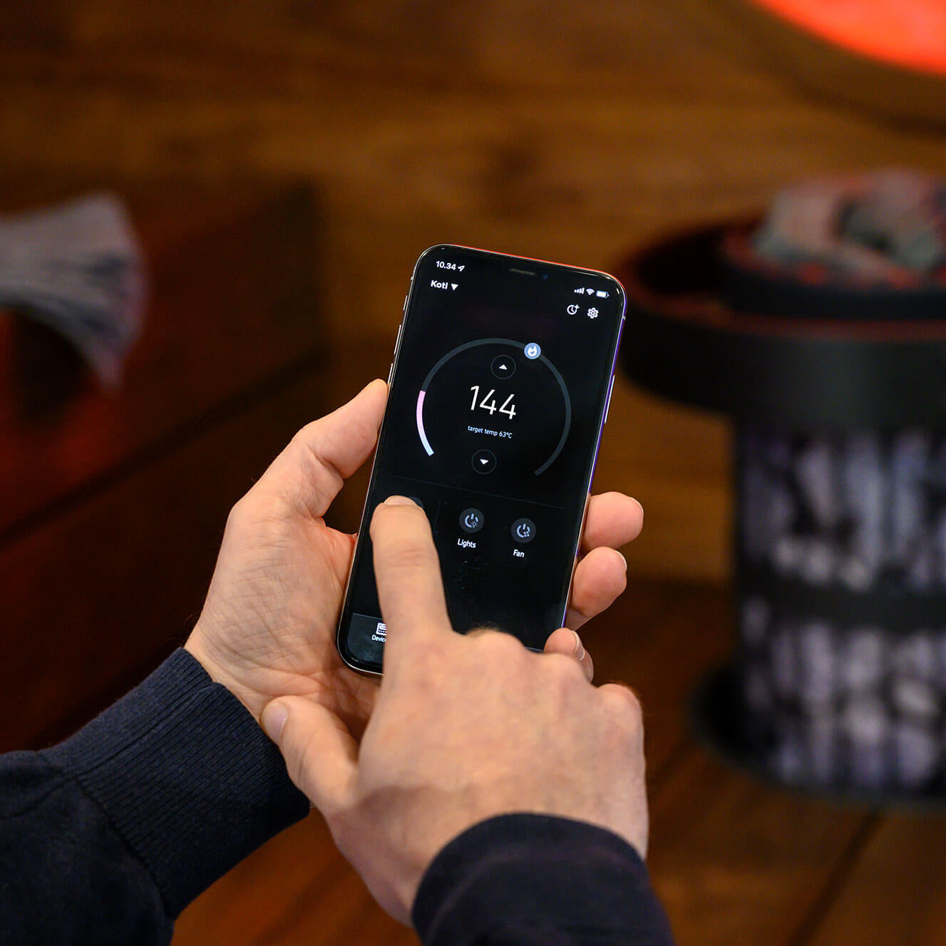 Person holding a smartphone with a screen displaying a measurement app, the Harvia MyApp, for remote control of Harvia heaters, on a wooden surface.