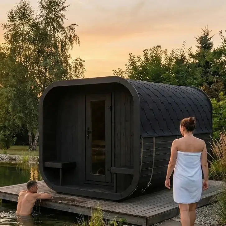 SaunaLife's CL12GCPB modern dark wooden outdoor sauna on a floating platform by a pond at sunset, with a woman wrapped in a white towel walking beside it and another person sitting in the water.