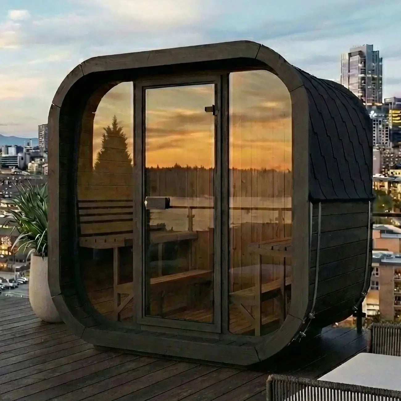 SaunaLife CL7GB, s modern outdoor sauna cabin with a dark wood exterior, large glass windows, and a curved front design, shown on a rooftop deck with a city skyline and sunset reflections.