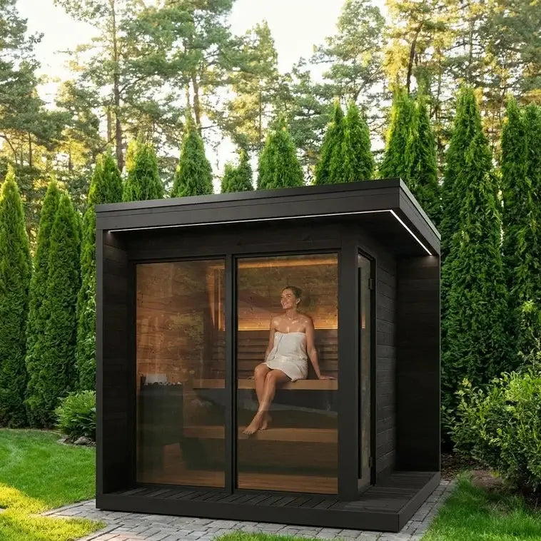 SaunaLife's modern GL6 black wooden outdoor sauna cabin with a large glass door and windows, shown in a landscaped garden with tall evergreen trees. A woman is seated inside on wooden benches, wearing a white dress.