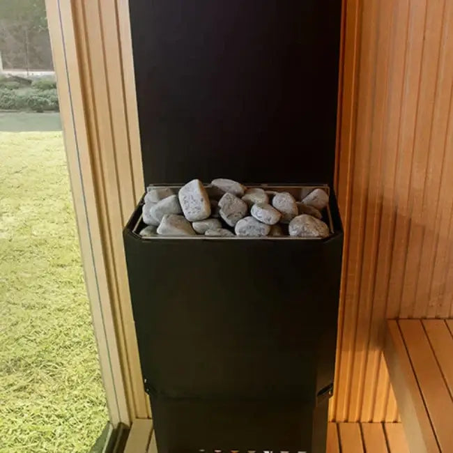 Saunum AIR 5 climate-equalizing electric sauna heater with stones inside, placed against a wooden wall with a window view.