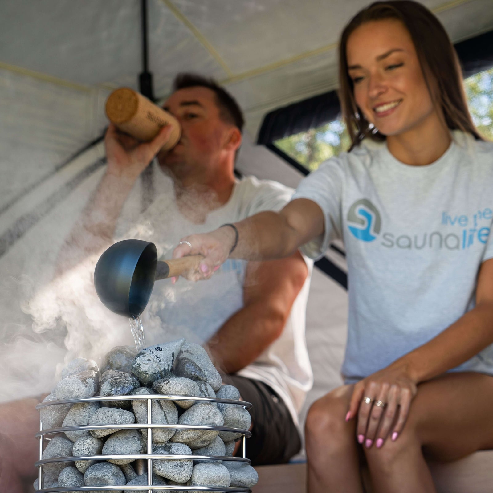 SaunaLife SaunaTrekker Portable Sauna Tent with adults enjoying sauna experience and pouring water on hot stones.