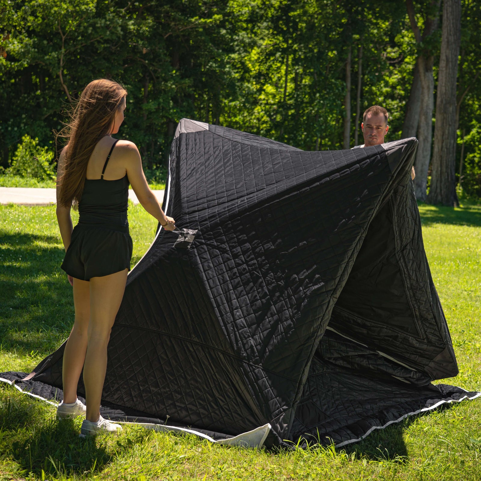 Two people setting up the SaunaLife SaunaTrekker Portable Sauna Tent in a green outdoor setting.