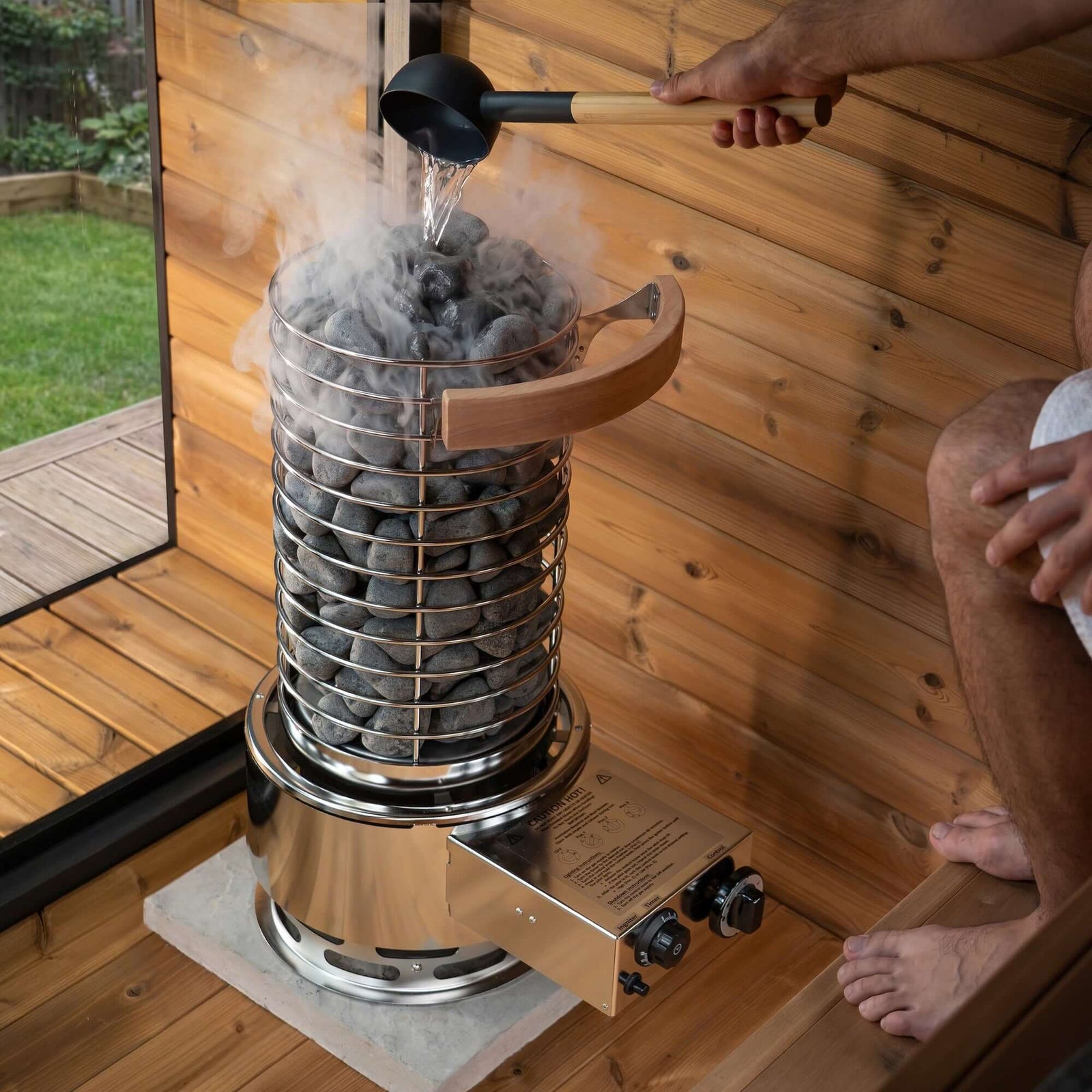 Person pouring water on sauna stones near SaunaLife TITAN SL1 Portable Propane Sauna Heater.