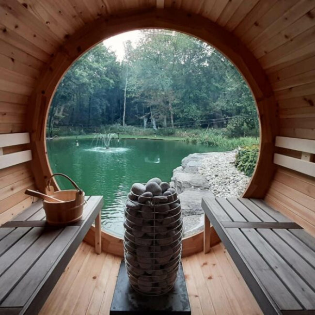 Huum Hive Mini sauna heater in wooden barrel sauna with a large circular window overlooking a serene lake and forest.