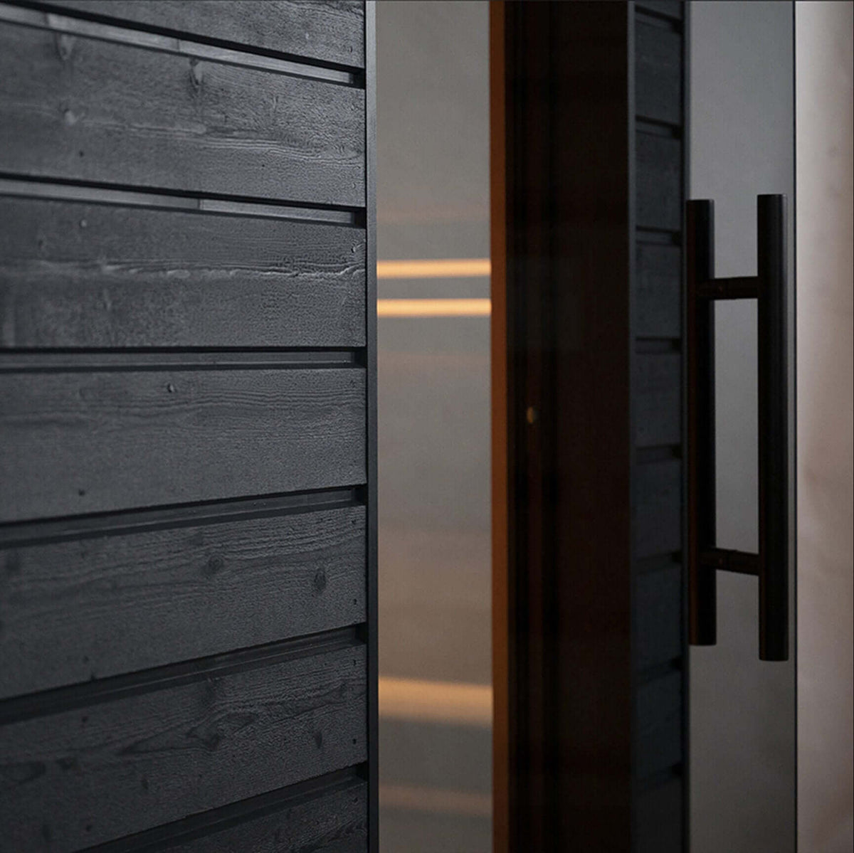 Close-up of a the modern exterior wall of SaunaLife's G6, with dark charcoal horizontal wood siding and a glass door with a black handle, showing warm interior light through the glass.