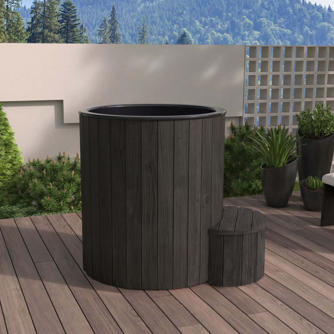 Black cylindrical S1B hot and cold plunge tub by SaunaLife, on a wooden deck with greenery and mountains in the background