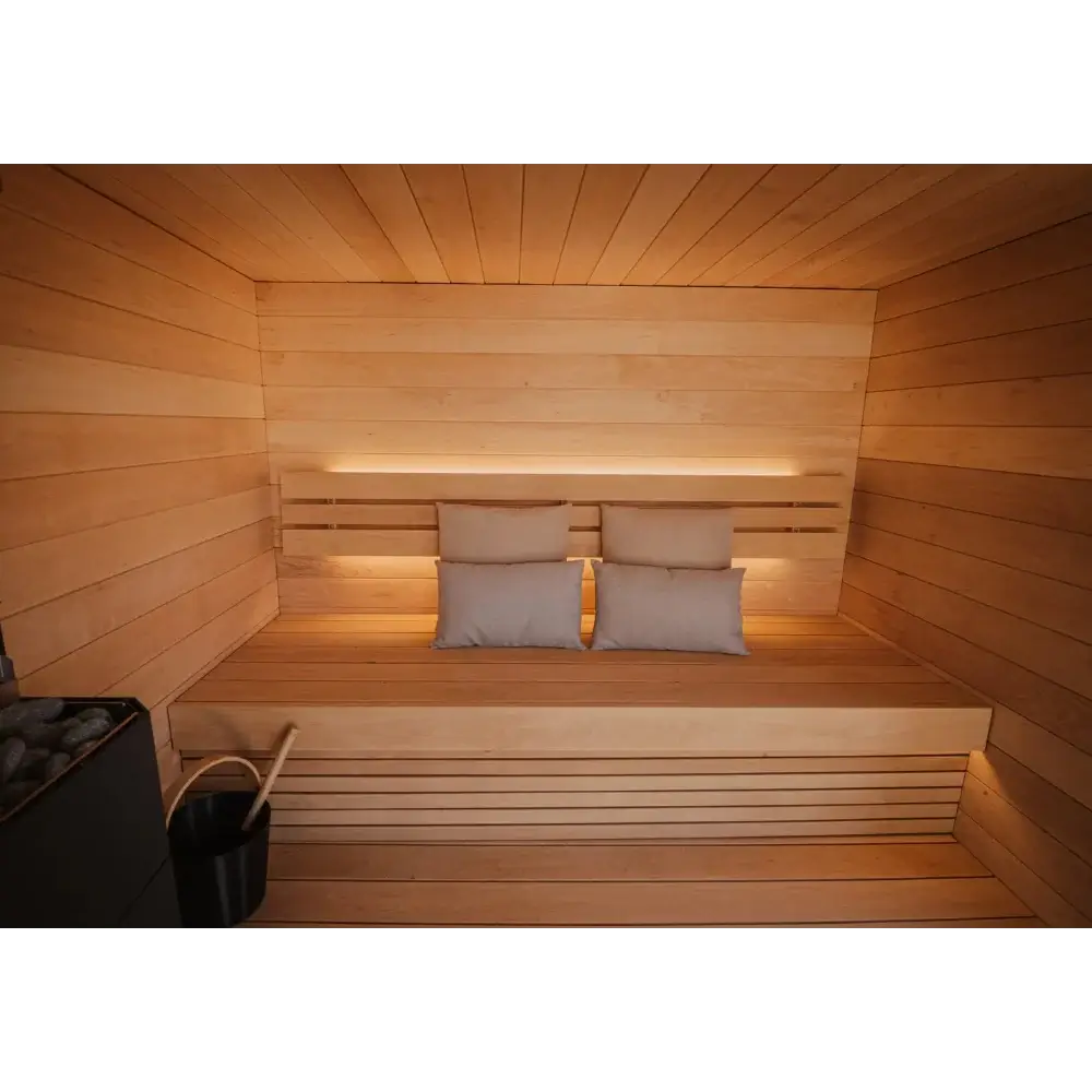 SaunaLife G6 wooden sauna interior with slatted wood walls and bench, warm ambient lighting, four beige pillows, and a black bucket with a wooden handle at the lower left as well as an electric heater.