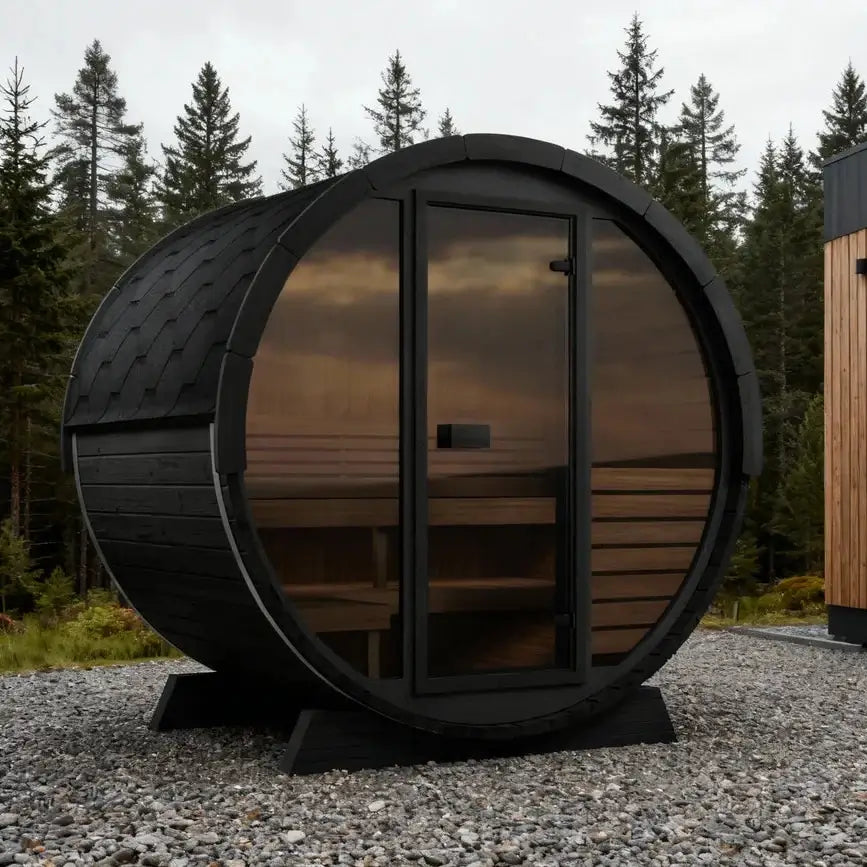 SaunaLife EE6G black wooden sauna cabin with large tinted glass door and windows stands on a gravel surface beside a modern wood-and-glass building, with a forest in the background.