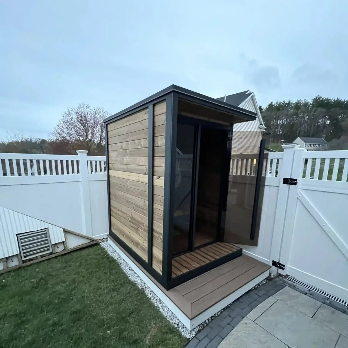 SaunaLife GL4, a modern outdoor sauna cabin with light wood paneling and a black metal frame, situated on a paved patio next to a white fence in a backyard setting.