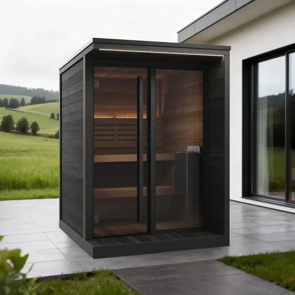 SaunaLife GL4B, a modern outdoor sauna cabin with dark gray wood-look exterior walls, a large glass door and window, set on a tiled patio beside a house with a green hillside landscape in the background.