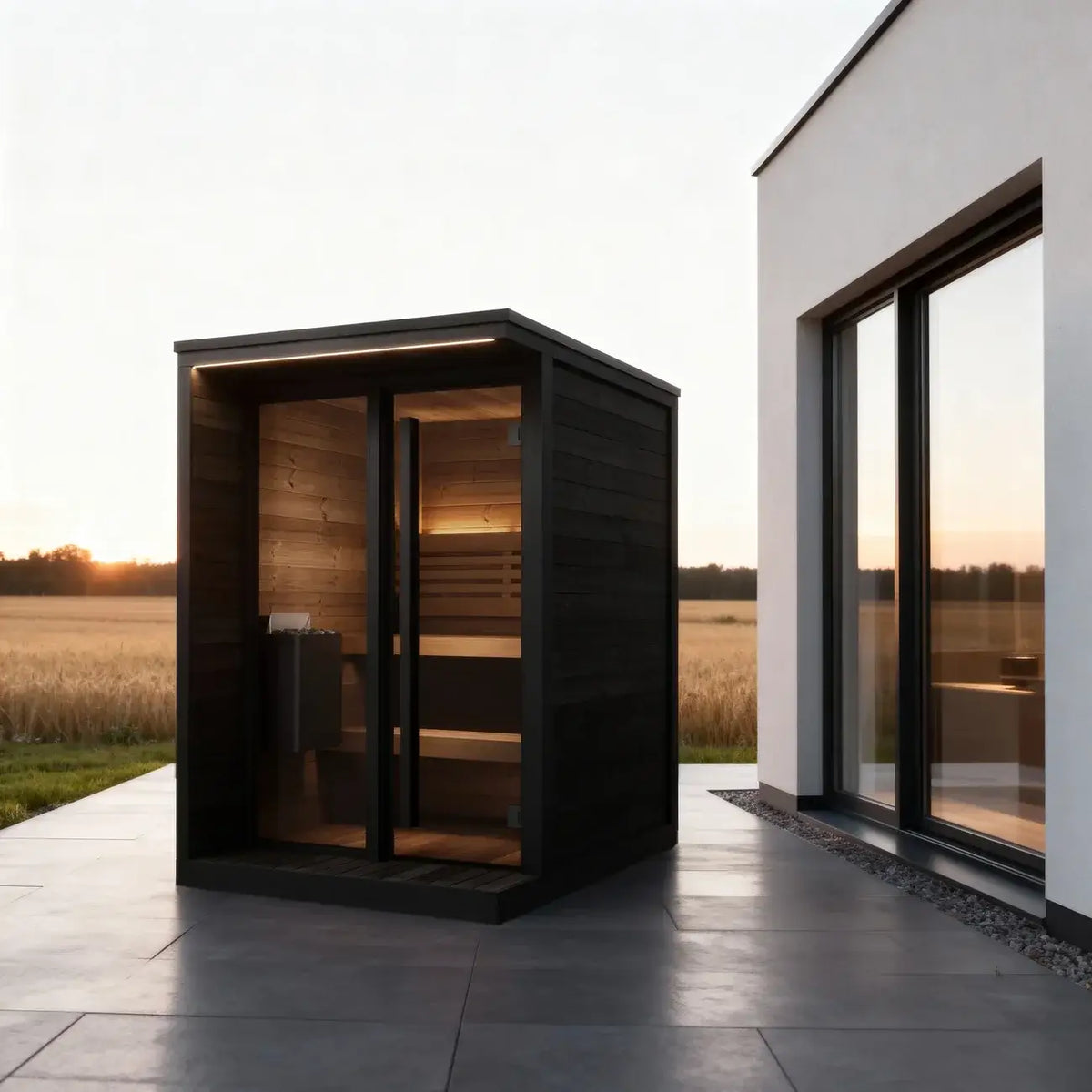 SaunaLife GL4B, a modern outdoor wooden sauna cabin with a dark black exterior and warm wood interior, situated on a tiled patio next to a white building with large windows during sunset.