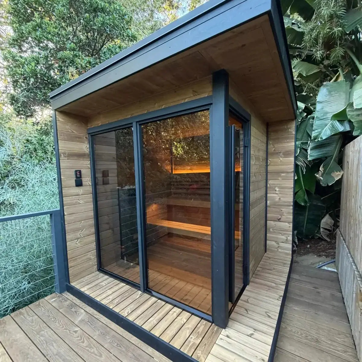 SaunaLife's GL6 modern wooden outdoor sauna cabin with a black metal frame, large glass windows and door, as well as a wooden deck, surrounded by lush green trees and plants.